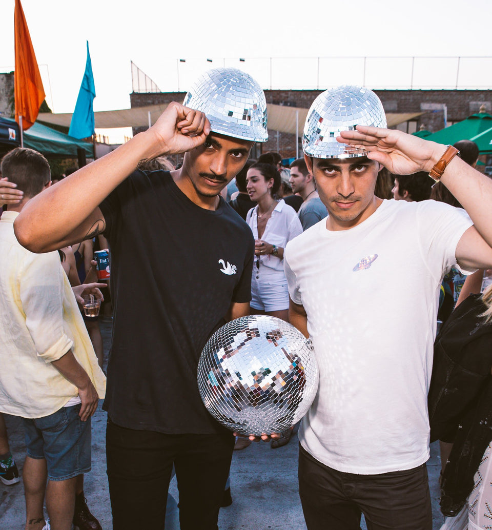 Two male models wearing the Thousand Deep disco hat, saluting towards the camera while holding a disco ball. Photo taken on rooftop at Lightning Society in Brooklyn, New York