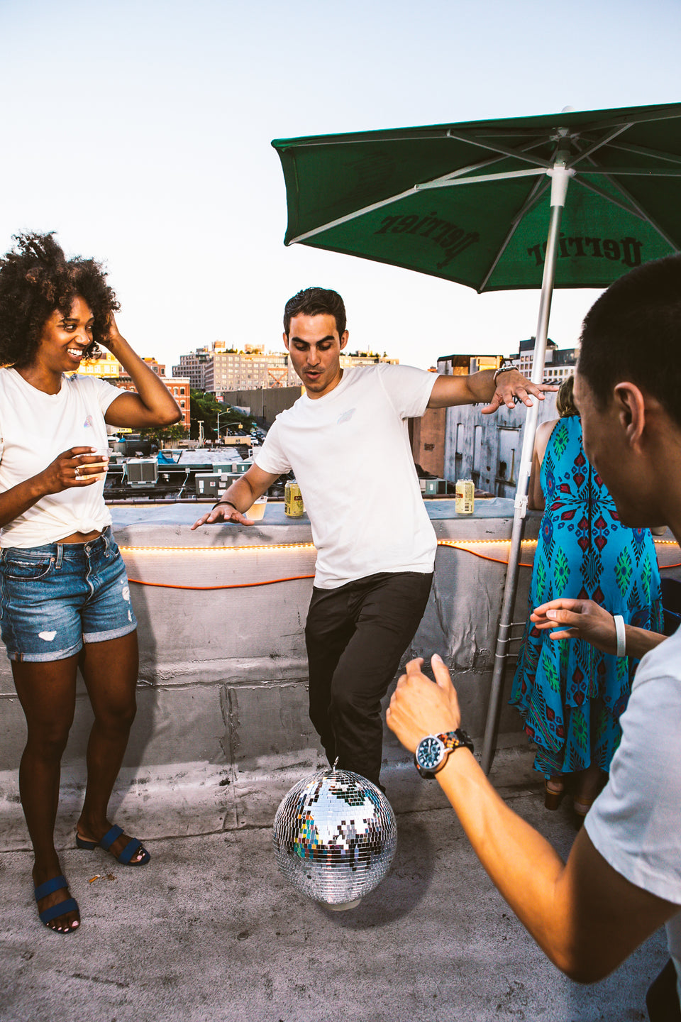 Male and female model wearing a Thousand Deep Planet Disco iridescent white shirt. Male model is balancing a disco ball on top of his foot while the female model watches in excitement. Photo taken on rooftop at Lightning Society in Brooklyn, New York
